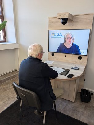 Eine Person sitzt am Tisch mit Unterlagen und Steuerungsmöglichkeiten gegenüber des Bildschirms, auf dem eine andere Person zu sehen ist.