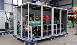 Eine große, modulare Wärmepumpenanlage in einer Werkhalle. Die Anlage ist in einem rechteckigen Metallrahmen verbaut, auf Rollen montiert und zeigt zahlreiche Rohre, Ventile und technische Bauteile, darunter ein grüner Kompressor und schwarze Druckbehälter. Im Hintergrund sind weitere Maschinen und Regale sichtbar